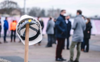 Spaten mit Helmen darauf steckt in Sand mit Personen dahinter
